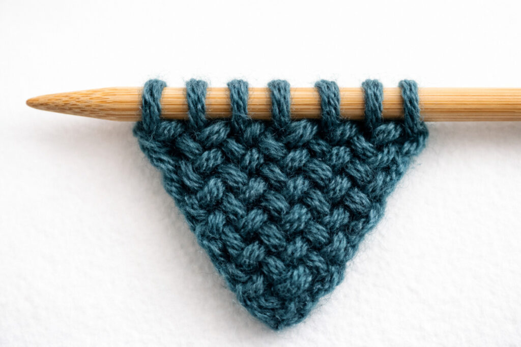 Close-up of seed stitch knitting on bamboo needles showing the narrow cast-on tip of a knitted tie in teal wool