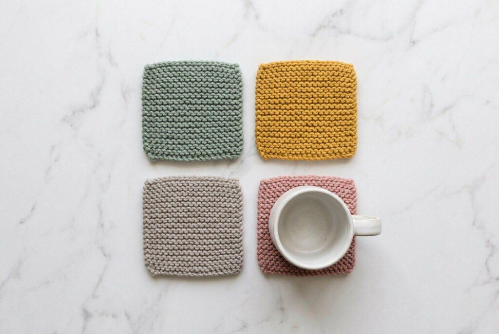 Four handknitted square coasters in mustard, sage green, pink, and grey on a marble surface with a white coffee mug