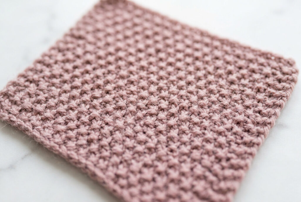Close-up of seed stitch knitted swatch in dusty rose yarn showing alternating knit and purl texture — reversible knitting stitch for scarves