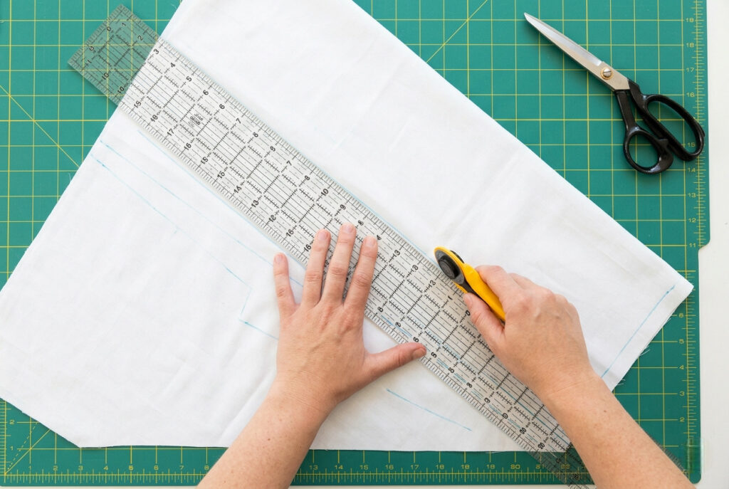 Hands cutting white cotton fabric on a cutting mat with a rotary cutter and ruler, with chalk measurement lines visible