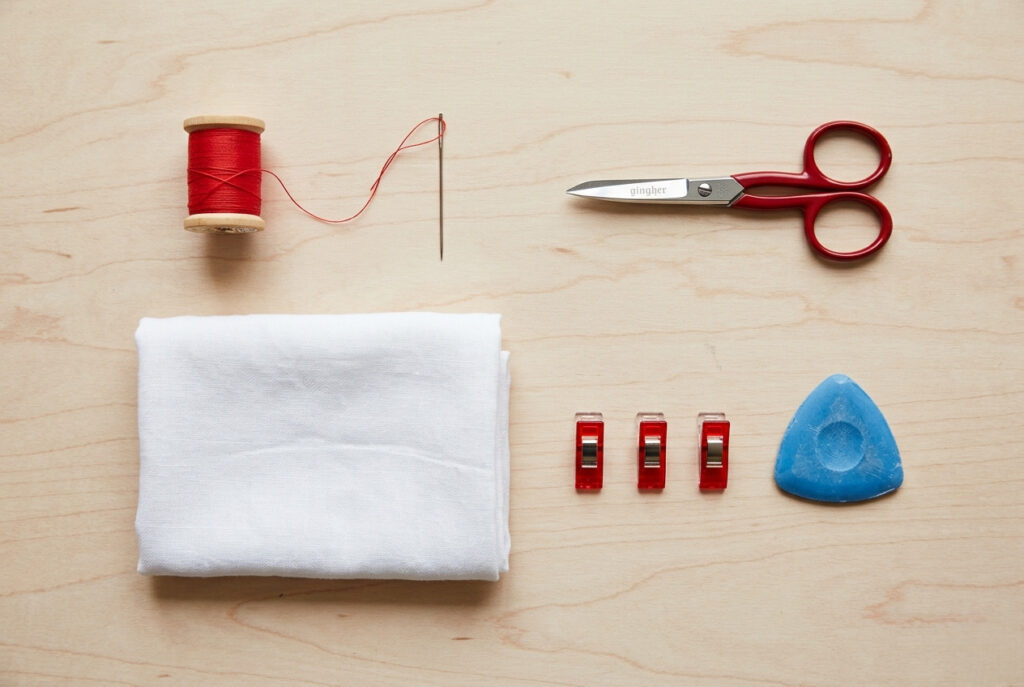 A flat lay of hand basting sewing supplies including a needle, contrasting red thread, fabric scissors, sewing clips, and fabric chalk on a wooden surface