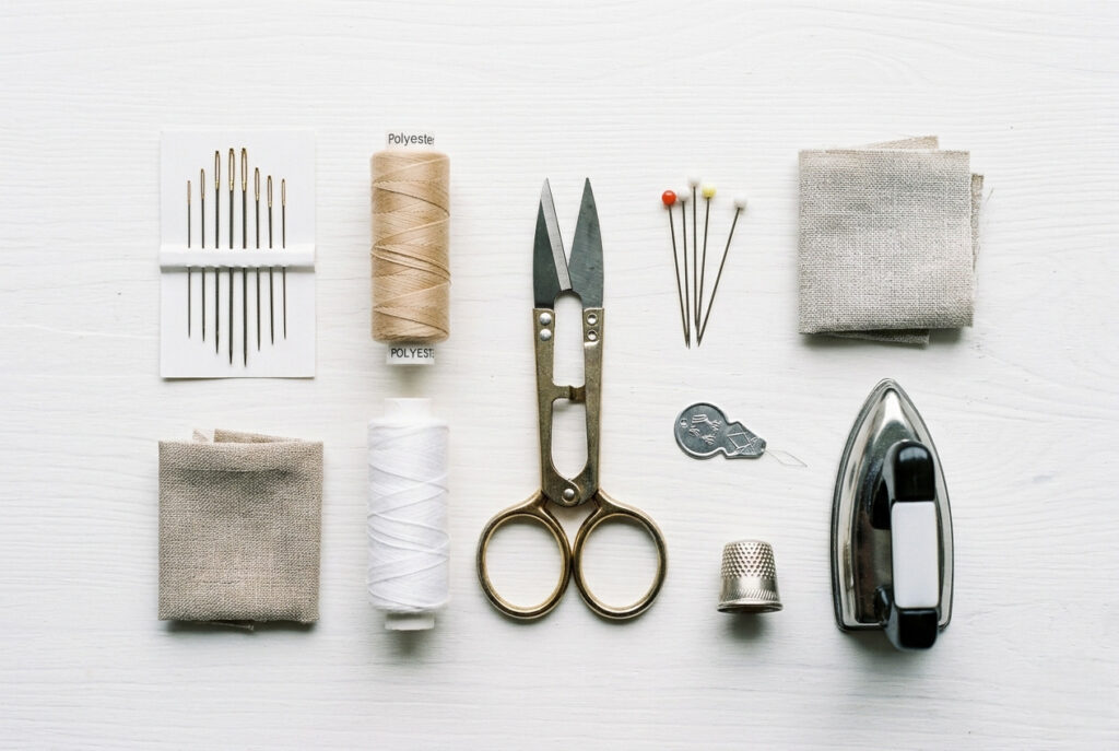 Flat lay of essential sewing repair tools including needles, thread spools, scissors, straight pins, a thimble, and a fabric patch on a white wooden surface