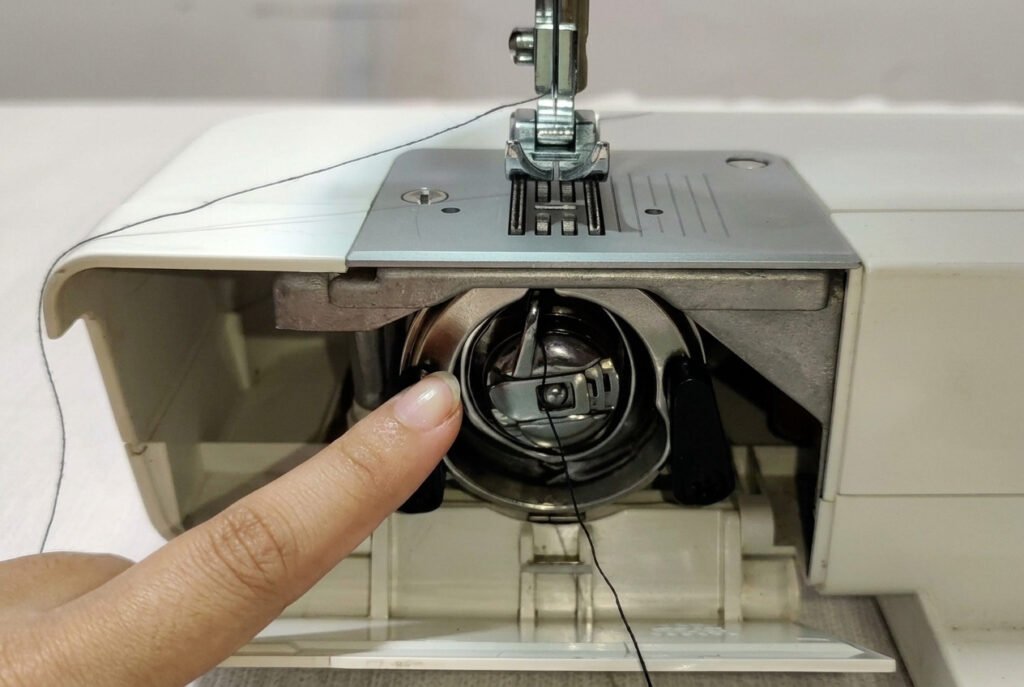 Finger pointing at the hook assembly while inspecting a sewing machine bobbin compartment