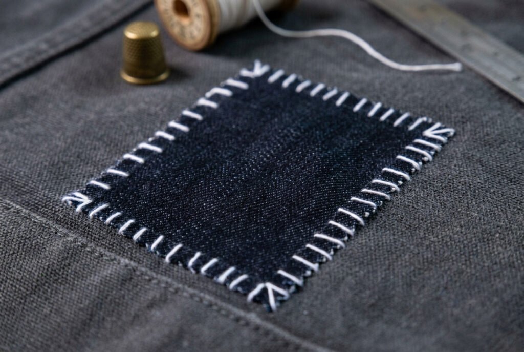 Denim patch whip-stitched onto gray canvas fabric, shown with a thimble and thread spool